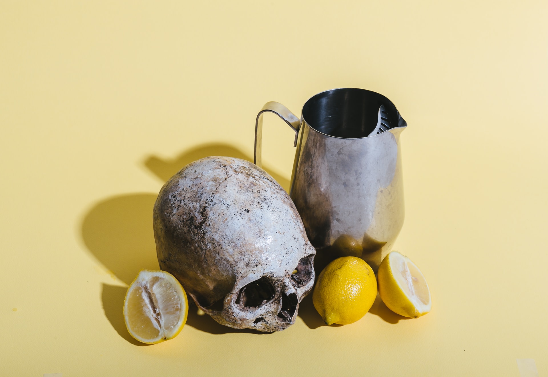 A skull and several lemons sit next to a metal pitcher in front of a yellow background.